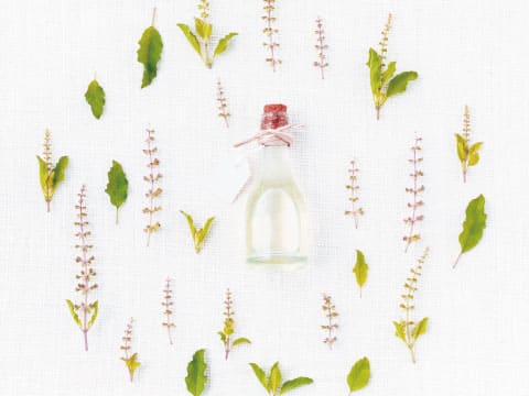 A small bottle of herbal essence sits at the center of leaves and small flowering sprigs arranged on a linen background.