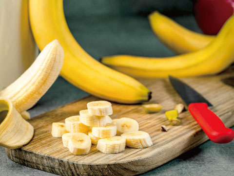 Fresh bananas sliced on a wooden cutting board with whole bananas and a red-handled knife nearby