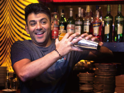 A smiling bartender shaking a metal Boston shaker in front of shelves lined with spirit bottles
