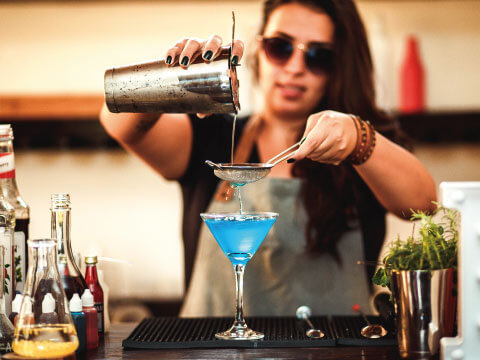 A bartender pouring a bright blue cocktail through a fine mesh strainer into a martini glass