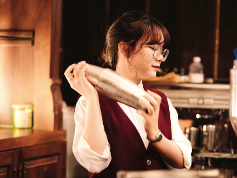 A bartender in a burgundy vest holding a cocktail shaker near her shoulder in a warmly lit bar