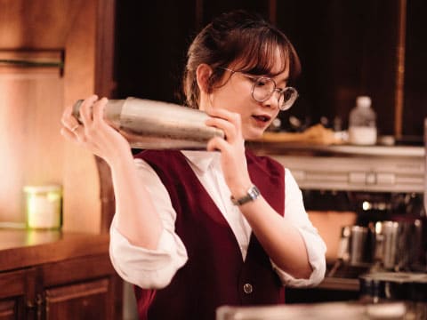 A bartender in a burgundy vest shaking a cocktail shaker over her shoulder in a dimly lit bar