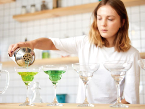 A bartender pouring a bright green cocktail from a cobbler shaker into a row of coupe glasses