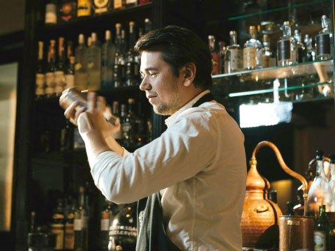 A bartender shaking a cocktail shaker in front of shelves lined with spirit bottles and a copper still