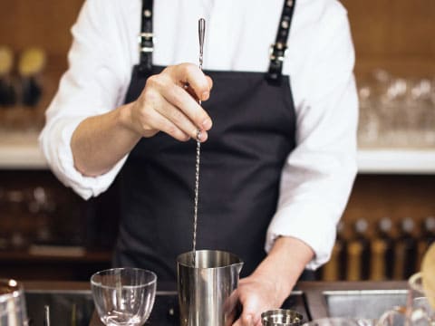 A bartender in a black apron stirring a cocktail in a mixing glass with a long bar spoon