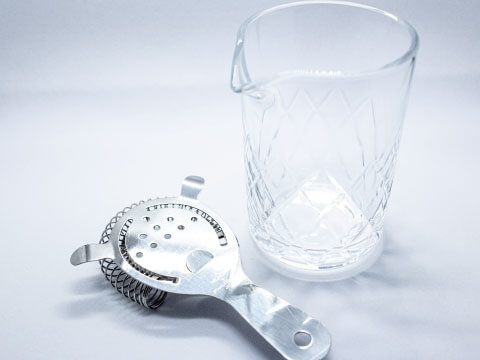 A cut-crystal mixing glass beside a stainless steel Hawthorne strainer with its coiled spring, laid on a white surface