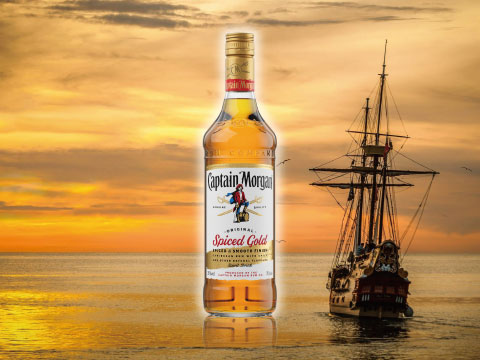A bottle of Captain Morgan Spiced Gold rum standing on the ocean at sunset, with a tall ship sailing in the background
