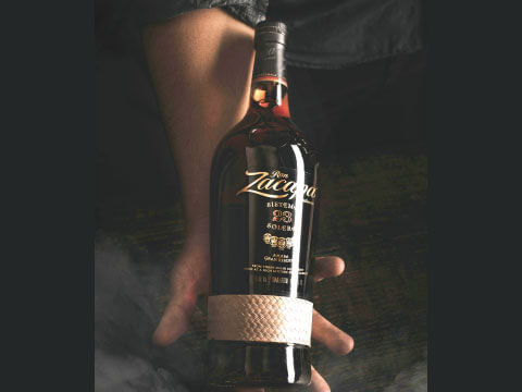 A hand holding a bottle of Ron Zacapa dark rum, wrapped in its signature woven band, against a dark moody background
