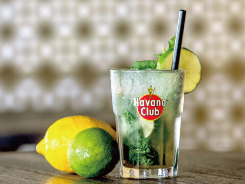 A mojito cocktail with mint and lime garnish in a Havana Club branded glass, beside a fresh lemon and lime
