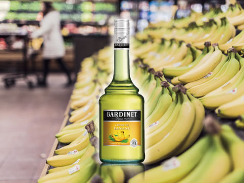 A bottle of Bardinet crème de banane liqueur standing among rows of fresh bananas at a grocery store
