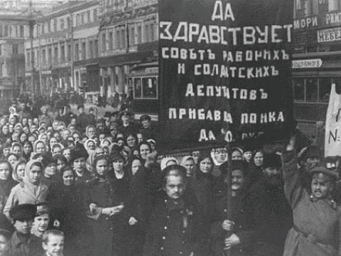 Historic black and white photo of a crowd marching in a Russian street during the 1917 Revolution, carrying a banner and Russian text sign