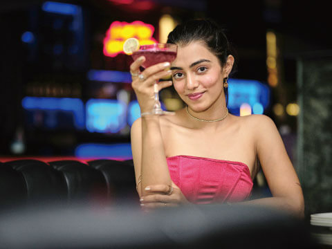 A woman in a pink dress holding up a red cocktail garnished with a lime wheel, seated at a bar with colorful lights in the background