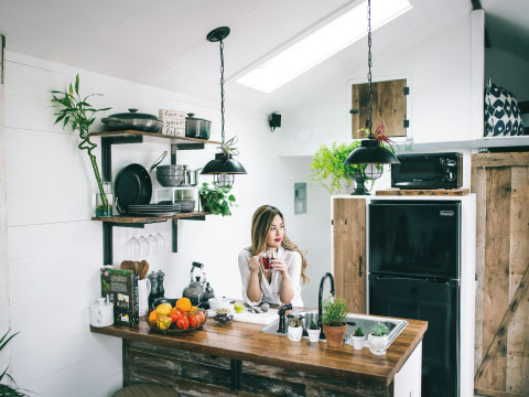 A woman sipping a cocktail at a rustic kitchen counter, surrounded by fresh fruit and greenery, representing home cocktail-making