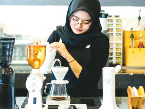 A barista wearing a hijab carefully prepares pour-over coffee using a dripper and kettle at a café counter.
