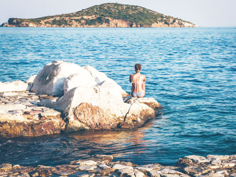 A person sitting on coastal rocks looking out at the ocean, with a small green island visible in the distance