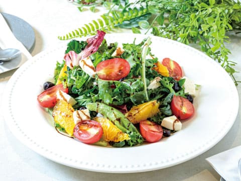 A white plate of citrus salad with orange segments, cherry tomatoes, black olives, feta cheese, and mixed greens