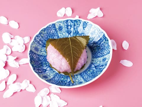 A pink sakura mochi wrapped in a preserved cherry leaf, served on a blue and white patterned dish with scattered cherry blossom petals