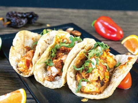 Three fish tacos topped with fresh cilantro, served on a black plate with lime and orange wedges and a red pepper