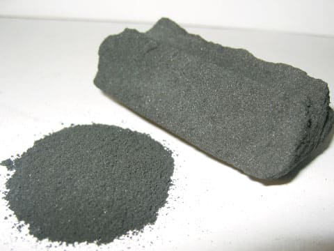 A solid block of activated charcoal beside a pile of fine activated charcoal powder on a white surface