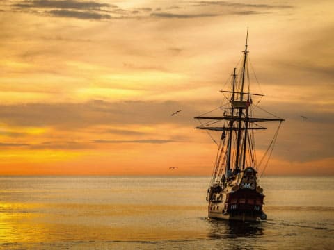A historic tall ship sails on calm ocean waters beneath a golden sunset sky with birds flying overhead.