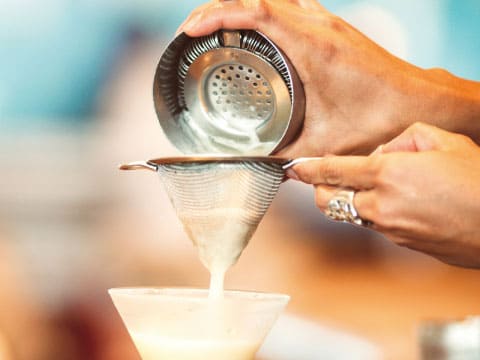 A bartender pours a creamy cocktail through a Hawthorne strainer and fine mesh strainer into a coupe glass.