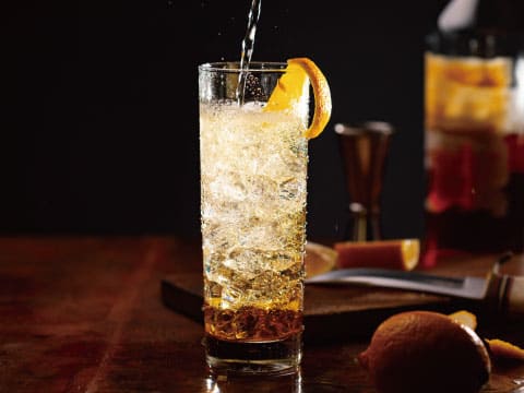 Soda water being poured into an amber highball cocktail garnished with an orange twist, with fresh oranges and bar tools nearby