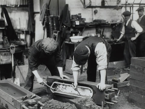 A black and white vintage photo of workers in flat caps using hand tools at a workbench inside a workshop