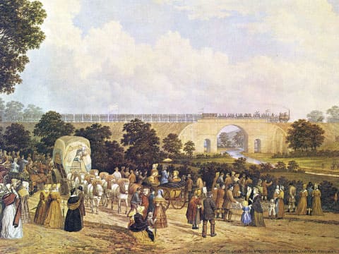 A historical painting shows crowds gathering to watch the first train cross a viaduct on the Stockton and Darlington Railway.