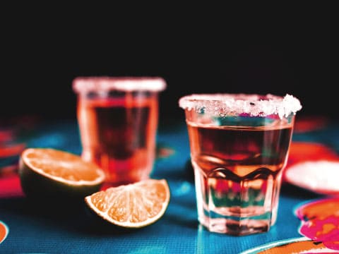 Two salt-rimmed tequila shot glasses sit on a colorful table beside cut lime wedges.
