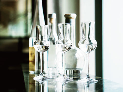 Three glasses of clear spirits stand on a bar counter beside bottles, with sunlight streaming through a window.