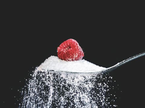 A single raspberry sits atop a heaping spoonful of sugar against a dark background.