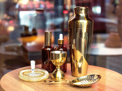 A gold-toned Cobbler shaker, Hawthorne strainer, jigger, and dropper bottles arranged on a wooden table