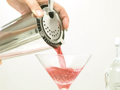 A hand holding a Hawthorne strainer over a shaker tin, pouring a red cocktail into a martini glass