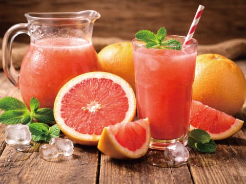 A pitcher and glass of fresh pink grapefruit juice garnished with mint, surrounded by cut grapefruit and ice on a wooden table