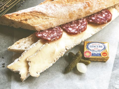 Sliced baguette layered with soft cheese and salami, served with cornichons and pearl onions on parchment paper