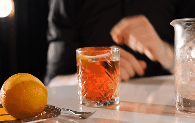 A bartender expresses citrus zest over an Old Fashioned cocktail on a bar counter, with a whole orange nearby.