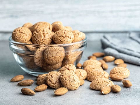 A glass bowl filled with Amaretti cookies, surrounded by scattered whole almonds on a gray surface