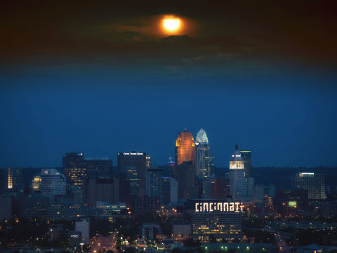 The illuminated downtown Cincinnati skyline at night, with a glowing moon rising above the skyscrapers and the city's name lit up on a building