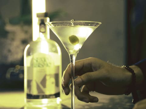 Hand holding a vodka martini glass garnished with an olive, vodka bottle blurred in background