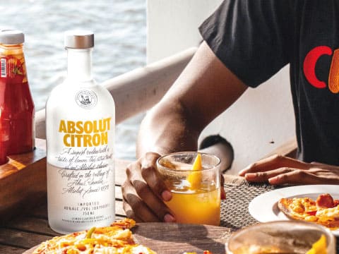Absolut Citron vodka bottle on an outdoor table with a citrus cocktail, pizza, and ketchup nearby