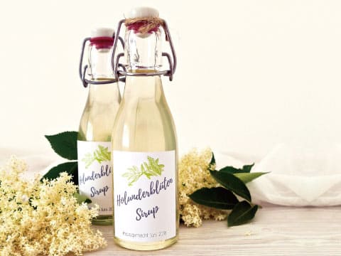 Two labeled bottles of elderflower syrup stand beside fresh elderflower blossoms and leaves.