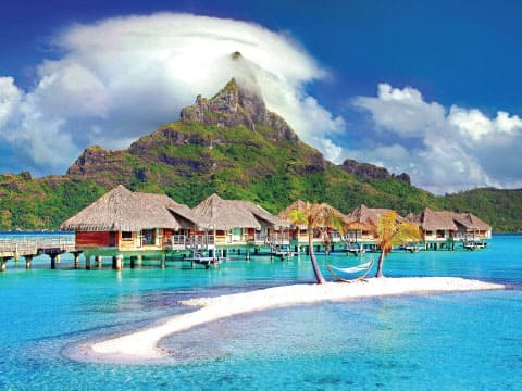 Thatched-roof overwater bungalows on stilts surrounding a small sandy island, with a dramatic mountain peak and turquoise water