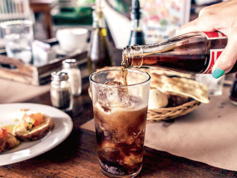 A hand pouring cola from a bottle into a glass of ice at a rustic café table with bread and small plates nearby