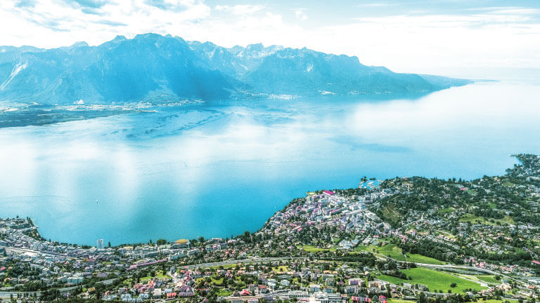 An aerial view of turquoise Lake Geneva surrounded by mountains, with lakeside towns dotting the shoreline
