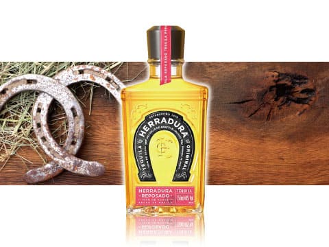 A bottle of Herradura Reposado tequila displayed between rustic horseshoes and straw on the left and a wooden surface on the right