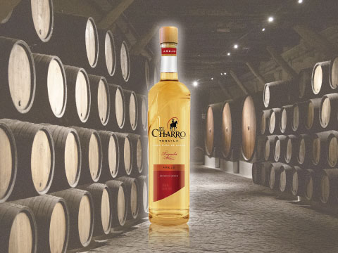 A bottle of El Charro Añejo tequila standing in a dimly lit cellar lined with rows of wooden aging barrels
