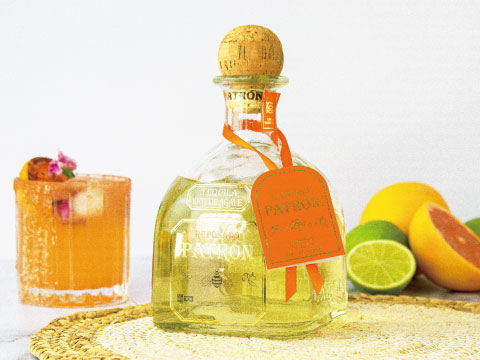 A bottle of Patrón tequila beside an orange cocktail garnished with flowers, surrounded by fresh citrus fruits on a woven placemat