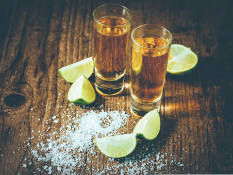 Two amber tequila shots served on a wooden table with fresh lime wedges and a pile of coarse salt