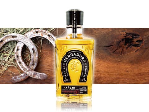 A bottle of Herradura Añejo tequila displayed between rustic horseshoes and straw on the left and a wooden surface on the right