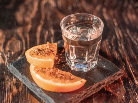 A clear tequila shot in a glass beside citrus slices coated with chili powder and salt, presented on a dark slate board with a rustic wood background